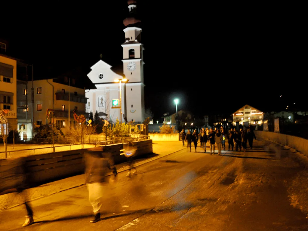 Beleuchtete Dorfkirche bei Nacht, davor eine Personengruppe.